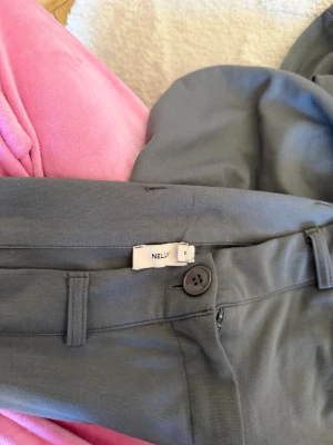 Grå kostymbyxor från Nelly - Snygga grå kostymbyxor från Nelly med vida ben och hög midja. Byxorna har bälteshällor, knapp och dragkedja framtill. Perfekta för en clean och stilren look. Materialet känns mjukt och följsamt.