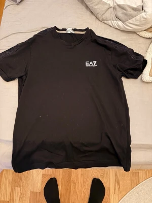 Svart EA7 t-shirt från Emporio Armani - Säljer en svart t-shirt från EA7 Emporio Armani med logga på bröstet. T-shirten har korta ärmar och en klassisk rund halsringning. Materialet är mjukt och bekvämt, perfekt för en avslappnad och stilren look.