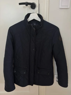 Marinblå quiltad jacka med hög krage - Snygg marinblå quiltad jacka från Stockh LM med hög krage och knappar framtill. Jackan har två fickor med knappstängning och diskret Barbour-logga på ärmen. Perfekt för dig som vill ha en stilren och tidlös look.