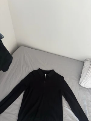 Svart långärmad tröja med dragkedja - En svart långärmad tröja med krage och kort dragkedja framtill. Tröjan har en slim fit passform och är tillverkad i ett mjukt och stretchigt material. Perfekt för dig som gillar en clean och stilren look.