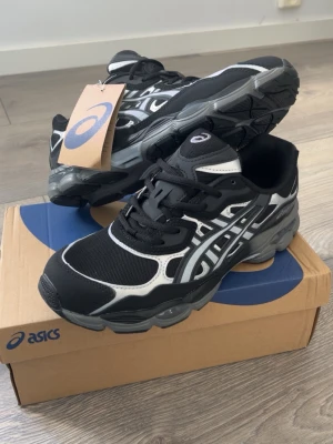 ASICS svart och vit sneakers - Svarta och vita ASICS sneakers med grå detaljer och klassisk mesh- och syntetöverdel. Skorna har en robust sula och ikoniska ASICS-loggan på sidan. Perfekt för dig som gillar sportig stil och komfort. Levereras i originalkartong.
