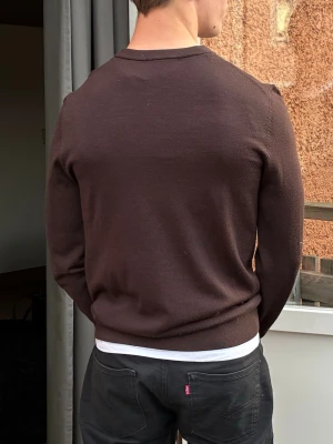 Brun stickad tröja - Mörkbrun stickad tröja med rund hals och långa ärmar. Tröjan har en enkel och stilren design utan mönster, med ribbade muddar vid ärmslut och nederkant. Perfekt att bära över en t-shirt för en avslappnad look.