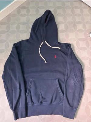 Marinblå hoodie från Polo Ralph Lauren - Snygg marinblå hoodie från Polo Ralph Lauren med klassisk huva och vit dragsko. Har en röd broderad logga på bröstet och en stor magficka. Perfekt för en avslappnad och stilren look.