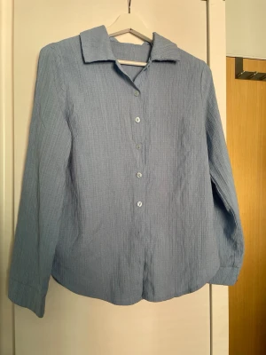 Ljusblå skjorta i bomull - En stilren ljusblå skjorta med lång ärm och klassisk krage. Skjortan har knäppning framtill med matchande knappar och är tillverkad i ett mjukt, lätt strukturerat bomullstyg. Perfekt för en avslappnad men snygg look.