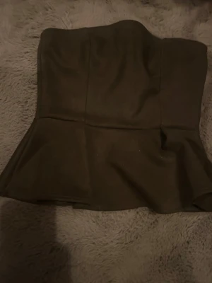 Brun axelbandslös peplum-topp - Snygg brun axelbandslös topp med peplum-utseende. Perfekt för att skapa en trendig och stilren look. Toppens form sitter tight upptill och släpper ut nertill för en feminin siluett.