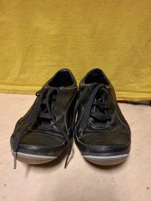 Svarta joggingskor från Puma - Säljer ett par svarta joggingskor från Puma i läder med klassisk låg profil och svarta snören. Skorna har vita detaljer på sulan och diskret Puma-logga på sidan. Perfekta för en avslappnad och sportig stil.