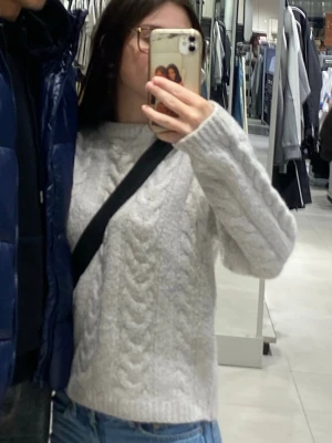 Ljusgrå kabelstickad tröja - Ljusgrå stickad tröja med klassiskt kabelstickat mönster och rund halsringning. Tröjan har långa ärmar och ribbade muddar vid ärmslut och nederkant. Perfekt för lager-på-lager och chill dagar.