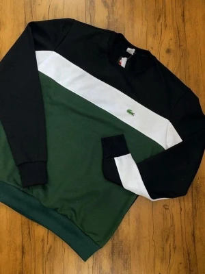 Lacoste sweatshirt i svart, grön och vit - Cool sweatshirt från Lacoste i svart, grön och vit med klassisk krokodillogga på bröstet. Tröjan har en bred vit diagonal rand över bröstet och ärmarna, samt ribbade muddar och rund hals. Perfekt för en avslappnad streetstyle-look.