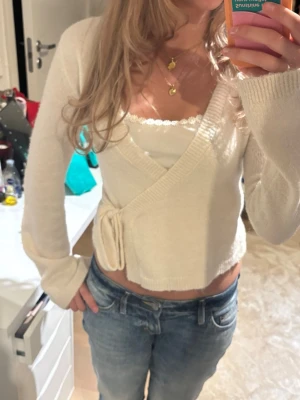 Krämvit omlottkofta med knytband - Söt krämvit omlottkofta med knytband i sidan och långa ärmar. Koftan har en croppad passform och är stickad i mjukt material, perfekt att slänga över ett linne eller topp. Enkel och stilren design som passar till jeans eller kjol.