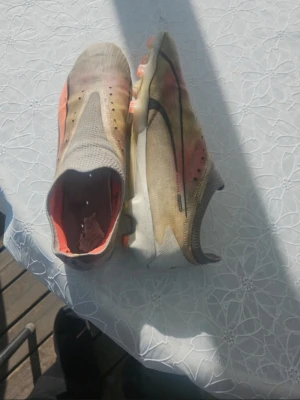 Nike fotbollsskor beige/orange - Säljer ett par Nike fotbollsskor i beige med orange och svarta detaljer. Skorna har låg profil, strömlinjeformad design och dobbar för bra grepp på planen. Ovandelen är i syntetmaterial och insidan har en röd/orange ton. Perfekta för dig som vill ha lätta och snabba skor.