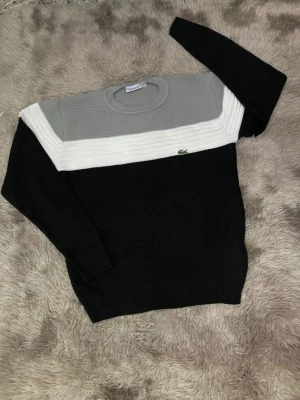 Lacoste tröja i svart, vitt och grått - Snygg stickad tröja från Lacoste med färgblock i svart, vitt och ljusgrått. Tröjan har rund halsringning, långa ärmar och den klassiska gröna krokodilloggan på bröstet. Perfekt för en stilren och avslappnad look.