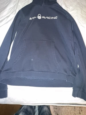 Mörkblå Sail Racing hoodie - Mörkblå hoodie från Sail Racing med vit logga tryckt på bröstet. Klassisk känguruficka framtill och huva. Perfekt för dig som gillar sportig och stilren design. Mjuk bomullskänsla och långa ärmar.