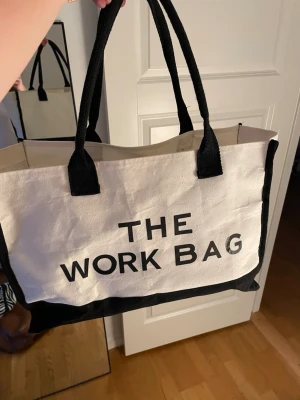 Vit och svart tygshoppingväska - Stor shoppingväska i vitt och svart tyg med texten 'THE WORK BAG' tryckt på framsidan. Väskan har två svarta handtag och rymligt huvudfack. Perfekt för att bära med sig allt man behöver till skolan eller jobbet.