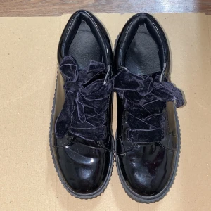 Svarta lackade sneakers med sammetssnören - Snygga svarta sneakers i glansigt lackmaterial med breda svarta sammetssnören. Skorna har en klassisk låg modell och en räfflad sula som ger extra edge. Perfekta för dig som vill sticka ut med en unik detalj på fötterna. De är som nya 