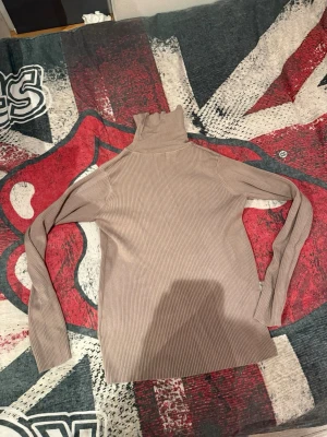 Beige ribbad polotröja - Säljer en beige ribbad polotröja med lång ärm och hög krage. Tröjan är figurnära och tillverkad i ett stretchigt material som sitter snyggt på kroppen. Perfekt att styla med jeans eller kjol för en clean look.