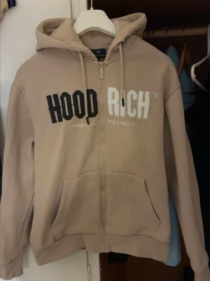 Beige ziphoodie från Hoodrich - Beige hoodie med dragkedja från Hoodrich. Stor huva, fickor framtill och tryck i svart och vitt på bröstet. Mjuk bomullskänsla och avslappnad passform, perfekt för en streetwear-look.