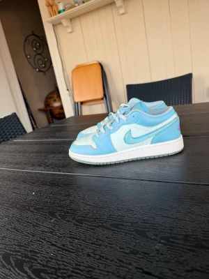 Nike Air Jordan 1 Low blå/vit - Snygga Nike Air Jordan 1 Low sneakers i ljusblått och vitt med rosa Air Jordan-logga på hälen. Skorna har klassisk låg siluett, vit sula och är tillverkade i en mix av läder och textil. Perfekta för dig som vill sticka ut med stilrena färger.