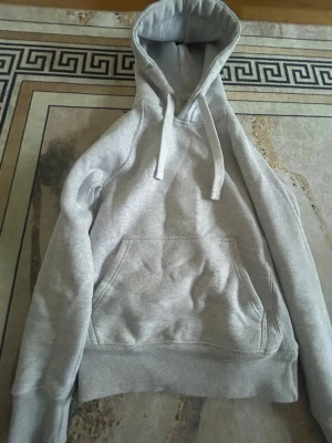 Ljusgrå hoodie med huva och ficka - En stilren ljusgrå hoodie med stor huva och dragsnören. Hoodien har en klassisk känguruficka framtill och ribbade muddar vid ärmslut och nederkant. Perfekt för en avslappnad look. Materialet känns mjukt och bekvämt, troligen bomullsblandning.
