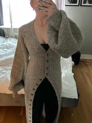 Adoore montana dress - Lång, ribbstickad grå kofta med stora ballongärmar och knappar hela vägen ner. Djup v-ringning och öppning framtill som ger en cool look. Perfekt att slänga över en outfit för extra värme och stil.