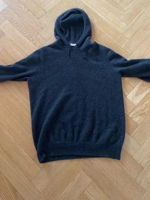 Mörkgrå Kashmir hoodie från Brown avenue - En enkel och stilren mörkgrå hoodie med huva och långa ärmar. Materialet är Kashmir och är mjukt och lite fluffigt, perfekt för att chilla eller hänga med kompisar. Ribbstickad nederkant och ärmslut ger en skön passform. 100% Kashmir nypris 3000kr 
