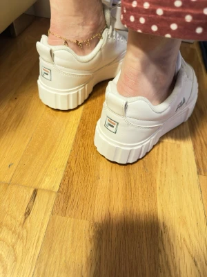Vita chunky sneakers från Fila - Säljer ett par vita chunky sneakers från Fila med höga, räfflade sulor och klassisk snörning. Skorna har Fila-logga på plös och häl, och är tillverkade i syntetmaterial med en modern, sportig look. Perfekta för dig som gillar streetstyle och vill sticka ut.