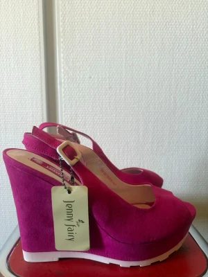 Cerise kilklackade pumps från Jenny Fairy - Snygga cerise pumps med kilklack från Jenny Fairy. Skorna har öppen tå, justerbar rem med vit spänne och är tillverkade i mockaimitation. Sulan är vit och ger en cool kontrast till den starka färgen. Perfekta för dig som vill sticka ut med din stil.