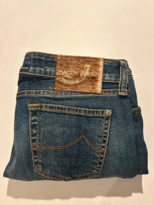 Blå jeans från Jacob Cohën - Klassiska blå jeans från Jacob Cohën med snygga kontrastsömmar och en unik patch i mocka med logga bak. Jeansen har fem fickor och rak passform, perfekta till din vardagsstil.