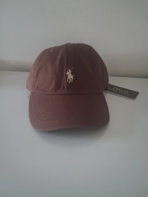Brun keps från Polo Ralph Lauren - Snygg brun keps från Polo Ralph Lauren med klassisk böjd skärm och broderad logga framtill. Justerbar rem baktill för perfekt passform. Tillverkad i mjuk bomull som ger en avslappnad och stilren look.