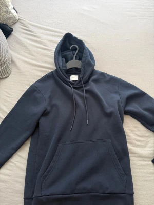 Mörkblå hoodie med ficka - Säljer en stilren mörkblå hoodie från Jack n Jones. Hoodien har en klassisk känguruficka framtill, dragsko i huvan och ribbade muddar vid ärmslut och nederkant. Perfekt för en chill och avslappnad look. Materialet känns mjukt och bekvämt.
