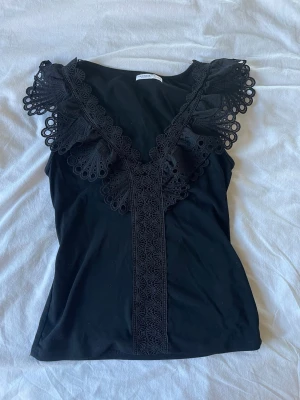 Svart spetsdetaljerad linnetopp - Snygg svart linnetopp med v-ringning och dekorativa spetsdetaljer längs ringningen och axlarna. Toppen är ärmlös och har en figurnära passform som framhäver siluetten. Perfekt att styla med jeans eller kjol för en trendig look.