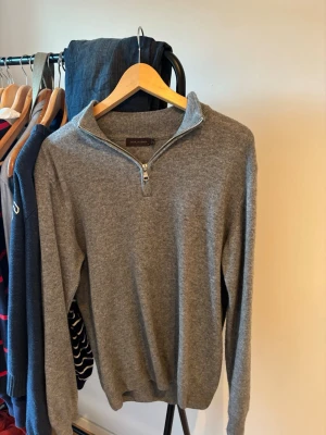 Grå halvzip-tröja från Oscar Jacobson - Klassisk grå tröja från Oscar Jacobson med halv dragkedja och ståkrage. Tillverkad i mjukt material, perfekt för lager-på-lager. Långärmad modell med ribbade muddar vid ärmslut och nederkant. Enkel och stilren design som funkar till många outfits.