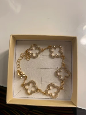 Guldfärgat armband med fyrklöver - Snyggt guldfärgat armband med flera fyrklöverformade länkar, dekorerade med små gnistrande stenar runt kanterna. Armbandet har justerbar kedja och smidig karbinhake. Perfekt accessoar för att lyfta din outfit.