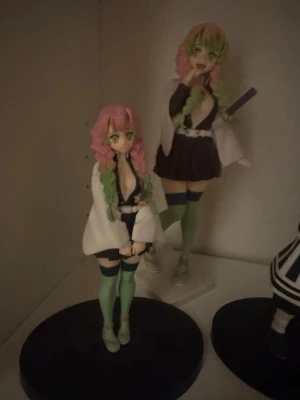 Demon Slayer Mitsuri Kanroji figurer - Två detaljerade animefigurer av Mitsuri Kanroji från Demon Slayer. Figurerna har rosa och grönt hår, vit haori, mörk klänning och gröna knästrumpor. Perfekt för samlare av anime eller manga-merch. Materialet är plast. kny