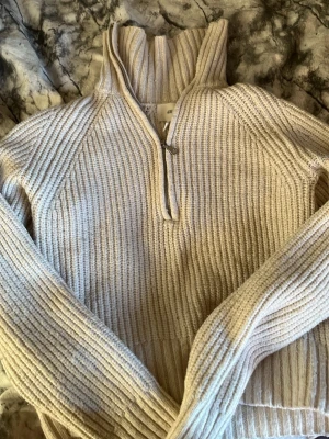 Beige stickad halvzip från H&M - Säljer en beige stickad tröja från H&M med halv dragkedja och hög krage. Tröjan har ribbstickat mönster och långa ärmar, perfekt för lager-på-lager. Enkel och stilren design som passar till det mesta.