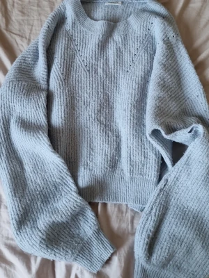 Ljusblå stickad tröja från Kappahl - Mysig ljusblå stickad tröja från Kappahl med breda ribbade muddar och diskret mönsterstickning framtill. Tröjan har rund halsringning och är gjord i ett mjukt, fluffigt material som känns skönt mot huden. Perfekt för kyliga dagar.
