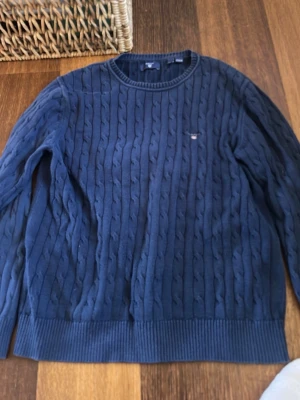 Kabelstickade tröjor från Gant - Två klassiska kabelstickade tröjor från Gant, en i marinblått och en i vitt. Båda har rund halsringning, ribbade muddar och diskret Gant-logga broderad på bröstet. Perfekta för en stilren och tidlös look. Materialet känns mjukt och bekvämt.