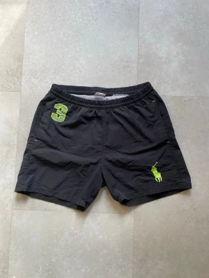 Svarta badshorts från Polo Ralph Lauren - Svarta badshorts från Polo Ralph Lauren med elastisk midja och gröna detaljer, inklusive en stor broderad logga och siffran 3 på framsidan. Tillverkade i ett lätt syntetmaterial, perfekta för sommaren.