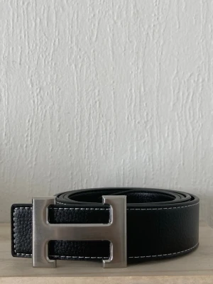 Svart skärp med H-spänne - Stilrent svart bälte i skinn med ett stort H-format metallspänne. Bältet har vit kontrastsöm längs kanterna och en klassisk design som passar till många olika outfits. Perfekt accessoar för att lyfta din look.