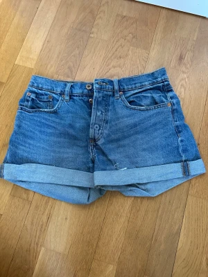 Blå denimshorts från 157 - Klassiska blå denimshorts från 157 med hög midja och uppvikta benslut. Shortsen har fem fickor, knappgylf och en avslappnad passform. Perfekta för varma dagar och enkla att matcha med olika toppar.