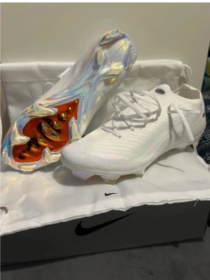 Vita Nike Mercurial fotbollsskor - Säljer ett par vita Nike Mercurial fotbollsskor med snygg, futuristisk design. Ovandelen är i lätt syntetmaterial och sulan har en skimrande, regnbågsfärgad finish med orange detaljer. Skorna har snörning och är perfekta för dig som vill sticka ut på planen.
