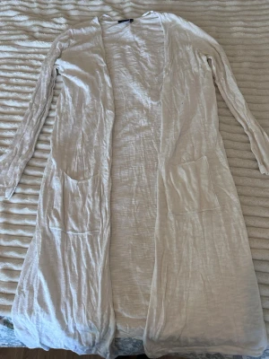 Lång beige kofta med fickor - Lång och tunn beige kofta med öppet fall och två stora fickor framtill. Koftan har långa ärmar och är tillverkad i ett mjukt, lätt material som ger en avslappnad look. Perfekt att slänga över en t-shirt eller topp för en chill stil.