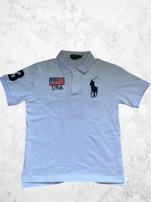 Vit piké från Polo Ralph Lauren - Vit pikétröja från Polo Ralph Lauren med USA-flagga och texten 'USA' på bröstet samt stor logga. Nummer 3 på ärmen och ryggen, samt 'UNITED STATES' och flagga på baksidan. Klassisk krage och korta ärmar. Perfekt för en sportig och stilren look.