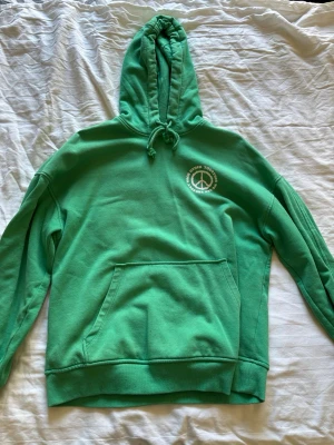 Grön hoodie med tryck och huva - Grön hoodie med stor vit text på ryggen: 'UNITY POWER LOVE' och ett peacemärke. På bröstet finns ett mindre tryck med peacemärke och text. Hoodien har huva med dragsko och en stor magficka. Perfekt för dig som gillar statement-plagg och sköna vardagslooks.