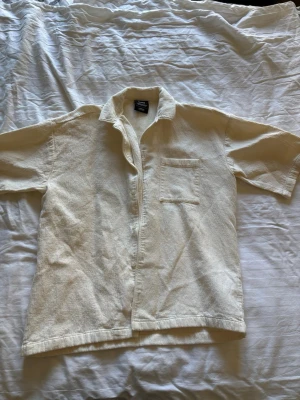 Benvit kortärmad skjorta i bomull - Säljer en beige kortärmad skjorta i mjuk bomull. Skjortan har klassisk krage, bröstficka och knappar framtill. Baksidan har ett tryck som ger en unik touch. Perfekt för dig som gillar enkel men snygg stil.