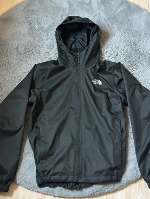 Svart vindjacka från The North Face - Snygg svart vindjacka från The North Face med huva och dragkedja framtill. Jackan har två sidofickor och elastiska muddar vid ärmslut och nederkant. Tillverkad i ett lätt och tåligt material som skyddar mot vind.