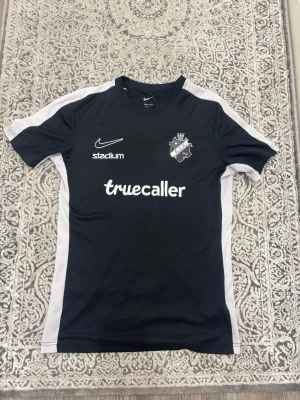 Svart AIK matchtröja från Nike - Svart AIK matchtröja från Nike med vita sidopaneler och tryckta loggor från Truecaller, Stadium, Fabege och SVEA. AIK:s klubbmärke på bröstet. Perfekt för fotbollsträning eller att bära på läktaren. Materialet är lätt och andas, designad för sport.