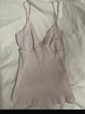 Ljusrosa ribbat  linne med spets - Sött ljusrosa linne med tunna axelband och ribbad struktur. V-ringad hals med spetsdetaljer längs kanterna ger en romantisk känsla. Perfekt för mysiga kvällar hemma.