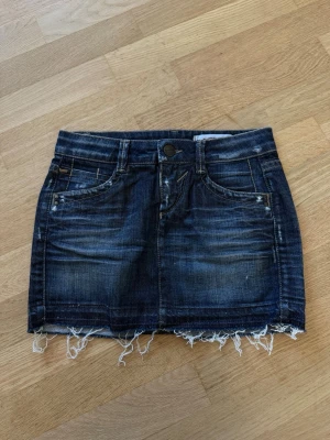 Mörkblå jeanskjol från GAS - Trendig mörkblå jeanskjol med slitna detaljer och fransig nederkant. Klassisk femficksmodell med knapp och dragkedja framtill. Perfekt för dig som gillar en avslappnad och cool stil. Bredden är 26cm så jag tänker att det blir storlek XS.