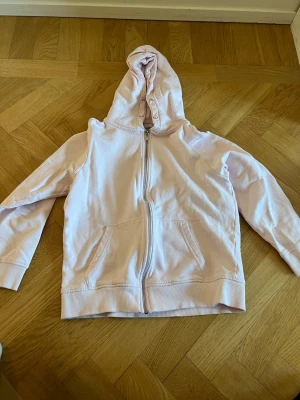 Ljusrosa hoodie med dragkedja - Säljer en ljusrosa hoodie med dragkedja framtill och två stora fickor. Hoodien har en klassisk huva och ribbade muddar vid ärmar och nederkant. Perfekt för en chill och avslappnad stil. Materialet känns mjukt och bekvämt.