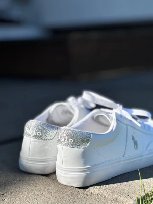 Vita Polo Ralph Lauren sneakers - Snygga vita sneakers från Polo Ralph Lauren med silverglittrig detalj på hälen och klassisk logga på sidan. Skorna har vita snören och är tillverkade i slätt konstläder för en clean look. Perfekta för dig som gillar stilrena och trendiga skor.
